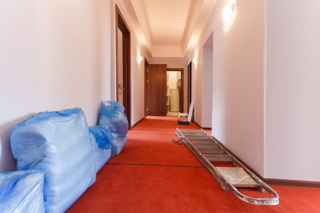 Corridor de l'hôtel pendant les travaux de rénovation, avec des objets enveloppés dans du plastique bleu le long du mur, une échelle en aluminium sur le tapis rouge et des portes de chambre en bois.