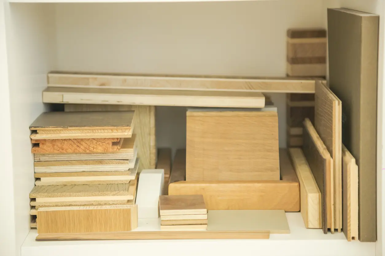 Texte altique Étagère blanche contenant des échantillons de bois empilés, notamment des blocs d’érable, des planches de bois synthétique, des chutes de contreplaqué et des panneaux stratifiés aux tons naturels.